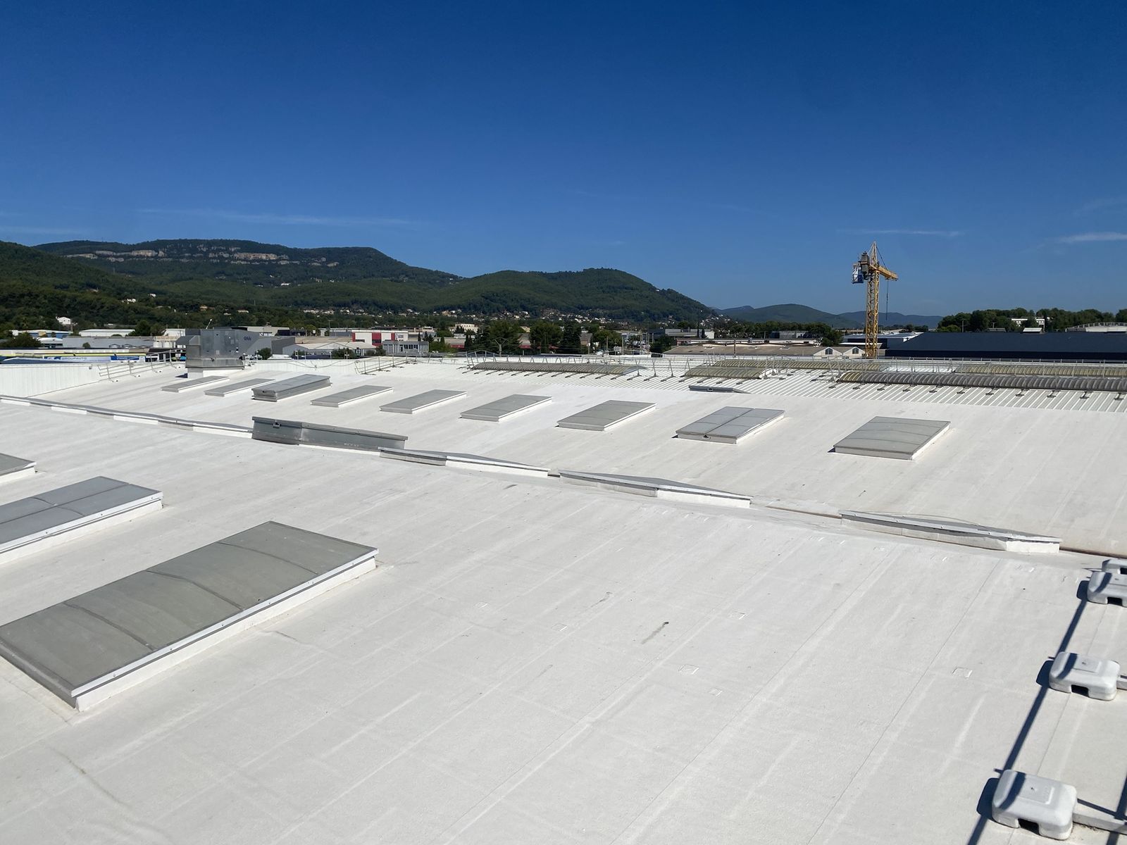 Grande toiture d'entrepôt peinte en blanc réflectif, avec ses lanterneaux, sous un ciel bleu, collines boisées au loin
