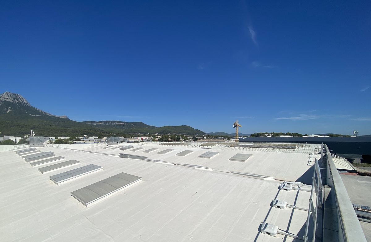 Vue large d'une toiture d'entrepôt entièrement blanche après application, lanterneaux et garde-corps en place, montagne au fond